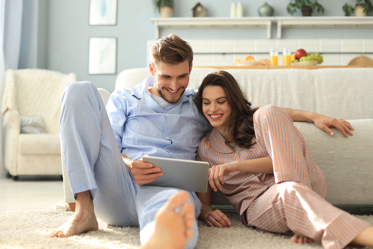 Ein Paar sitzt gemütlich im Pyjama auf dem Boden und schaut gemeinsam auf ein Tablet, umgeben von einem hellen Wohnraum.